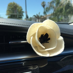 Puede incluir: Un ambientador para coche con forma de flor de fieltro color crema con centro negro, sujeto a una rejilla de ventilación del coche. La flor está enfocada, con el salpicadero y el exterior del coche de fondo.