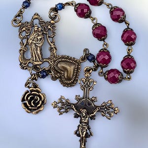 Puede incluir: Un rosario antiguo con cuentas facetadas de color rojo intenso y adornos de metal en tono bronce. El rosario presenta un crucifijo, un colgante en forma de corazón, un dije de rosa y un colgante con una figura religiosa.
