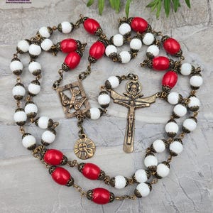Puede incluir: Un rosario con cuentas blancas y rojas, y detalles metálicos de color latón antiguo. El rosario presenta un crucifijo, una pieza central con una paloma y un colgante de sol. Las cuentas están ensartadas en una cadena con conectores decorativos.