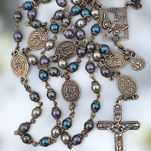 May include: A rosary with metallic beads in shades of gray, blue, and purple. The rosary features ornate metal pendants and a crucifix. The beads are strung together with a bronze-colored chain.