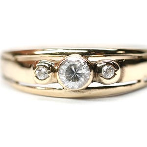 May include: A gold ring featuring a central round brilliant-cut clear stone flanked by two smaller round clear stones. The band has a split design with a polished finish, holding the three stones in a horizontal row across the centre of the band.