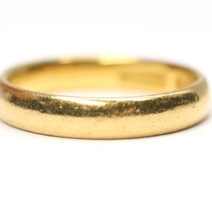 May include: A close-up shot of a gold wedding band. The ring is a solid, rounded band with a slightly worn appearance. The gold color is warm and lustrous, set against a clean white background. The ring's design is simple and classic.