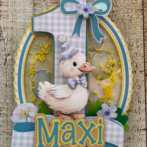 May include: A layered papercraft birthday decoration featuring a cartoon duck wearing a party hat and bow tie. The number "1" is prominent, with the name "Maxi" below. The design incorporates blue, yellow, and white gingham patterns and floral accents.