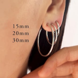 May include: Three silver-toned hoop earrings of varying sizes, labeled 15mm, 20mm, and 30mm. The hoops are thin, circular, and metallic, arranged in a nested formation within the earlobe. The text 15mm, 20mm, and 30mm is clearly visible.