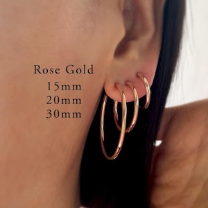 May include: Three rose gold hoop earrings in varying sizes, labeled as 15mm, 20mm, and 30mm. The hoops are thin, circular, and polished, worn in three separate piercings along the earlobe. The text Rose Gold 15mm 20mm 30mm is visible.