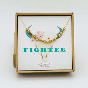 May include: A gold-colored beaded necklace with a delicate chain, presented in a small brown box. The box features a card with the word "FIGHTER" in teal, a floral bull skull graphic, and the text "secret message jewelry".
