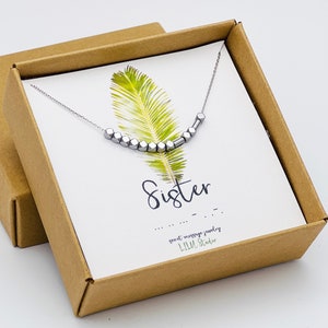 May include: A silver necklace with a message in braille that reads "Sister". The necklace is in a brown box with a green leaf design and a card that says "Sweet message jewelry, LILY Studio".