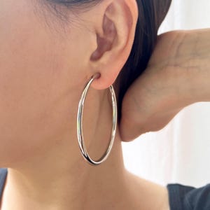 May include: A large, polished silver-tone hoop earring with a smooth, rounded surface. The earring is circular in shape and features a continuous, seamless design that hangs from the earlobe, reflecting light across its metallic finish.