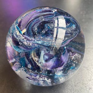 May include: A glass sphere with swirling patterns of blue, purple, and white. The sphere is filled with small air bubbles.