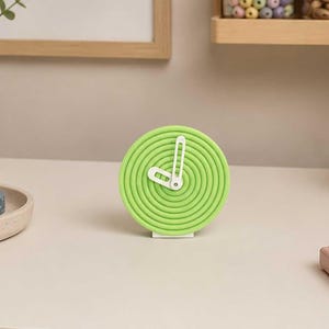 May include: A lime green desk clock with a white clock face and hands. The clock is circular with a spiral design. The desk also features washi tape, a plant, and a framed print that says "MAKE SOMETHING BEAUTIFUL."