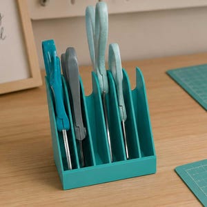 May include: A teal desk organizer holds scissors and pens. A framed print reads "create something beautiful." Rolls of decorative tape and a notebook with a floral cover are also visible. Two teal cutting mats and a metal ruler complete the craft supplies.