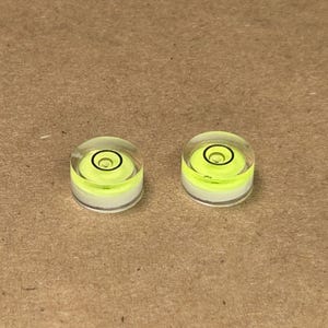 May include: Two small, clear, circular bubble levels with a bright yellow liquid and a black circular indicator. The levels are set on a light brown surface. These tools are used to determine if a surface is level.