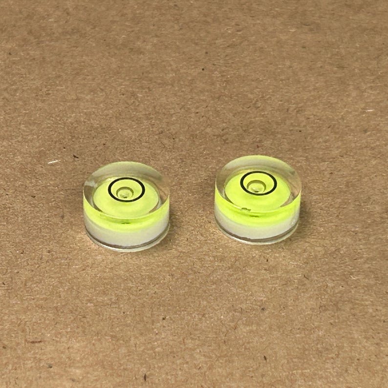Peut inclure: Deux petits niveaux &agrave; bulle circulaires et transparents, avec un liquide jaune vif et un indicateur circulaire noir. Les niveaux sont pos&eacute;s sur une surface marron clair. Ces outils servent &agrave; v&eacute;rifier le niveau d'une surface.