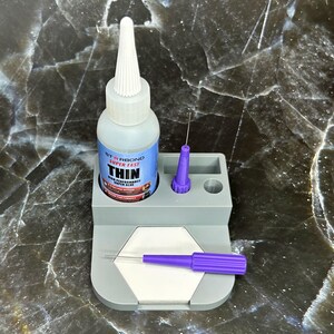 May include: A bottle of Starbond Super Fast Thin glue with a white applicator tip, set in a gray holder with a purple applicator and a needle. The holder has a white hexagonal surface. The background is a dark, marbled surface.