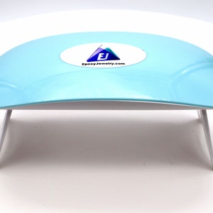 May include: A light blue UV lamp with a white base and two white legs. The lamp has a white oval sticker with the text "EJ EpoxyJewelry.com" on the top.