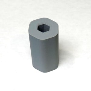 May include: A gray, hexagonal-shaped rubber roller with a hollow, six-sided center. The roller is cylindrical with rounded corners and a light gray rim around the top opening. The object is set against a white background.