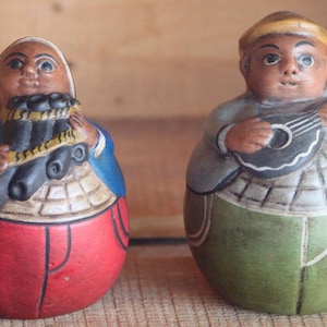 Puede incluir: Dos figuritas de cerámica, una tocando un instrumento musical y la otra sosteniendo un juego de pipas. La figurita que toca el instrumento lleva un atuendo verde y azul, mientras que la otra figurita lleva un atuendo rojo y azul.