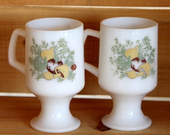 May include: Two white ceramic mugs with a footed base. The mugs have a green, brown, and yellow floral design with mushrooms and onions.