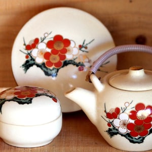 May include: A set of three ceramic teacups and a teapot with a floral design. The teapot has a light purple handle and a lid. The cups and teapot are decorated with white, red, and gold flowers.