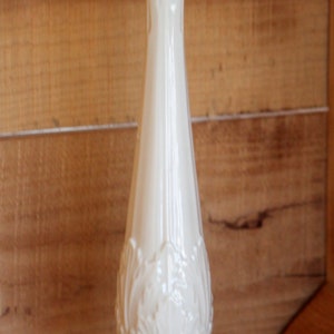 May include: A white ceramic vase with an ornate design. The vase has a tall, slender body and a wide base.