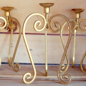 Puede incluir: Tres candelabros de metal dorado con un diseño decorativo en forma de corazón. Cada candelabro tiene una parte superior redonda y una base con un patrón de espiral.