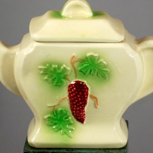 May include: A small, cream-coloured teapot with a lid, featuring a raised design of green leaves and a red berry. The teapot has a rounded body and a spout on one side.