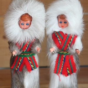 May include: Two dolls wearing traditional clothing and fur hats. The dolls have red and white patterned dresses with green accents. The dolls are standing on wooden bases.