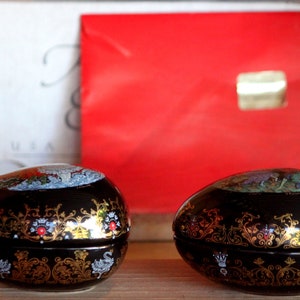 May include: Two black and gold egg-shaped trinket boxes with intricate floral designs. The boxes are closed and have a glossy finish.