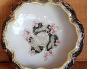 May include: A small, white porcelain bowl with two yellow handles. The bowl has a scalloped edge and is decorated with a floral design in pink, green, and gold.