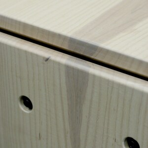 May include: Close-up of a light wood box with two black metal hinges and two black metal knobs. The wood grain is visible and the box has a smooth finish.