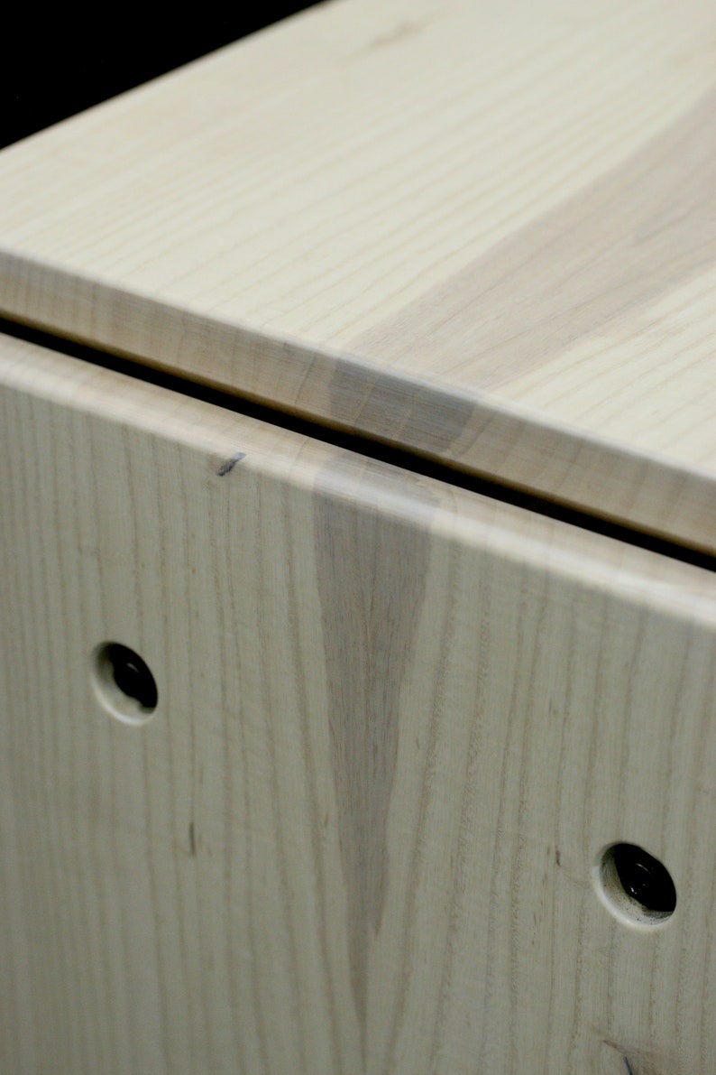 May include: Close-up of a light wood box with two black metal hinges and two black metal knobs. The wood grain is visible and the box has a smooth finish.