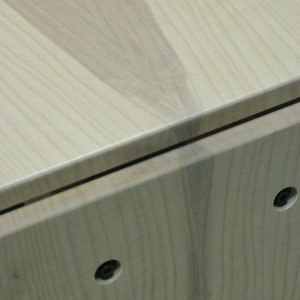 May include: Close-up of a light wood cabinet with a visible grain pattern. The cabinet has two small black screws on the front.