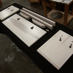 May include: A set of unfinished wooden pieces for a furniture project. The pieces are made of light-colored wood and have a natural grain pattern. The pieces are laid out on a black surface.