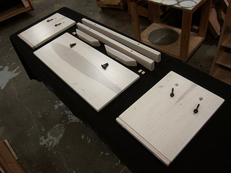 May include: A set of unfinished wooden pieces for a furniture project. The pieces are made of light-colored wood and have a natural grain pattern. The pieces are laid out on a black surface.