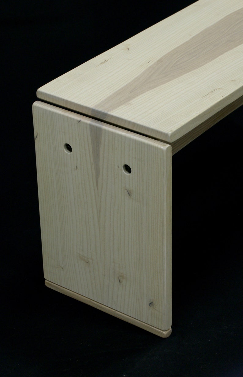 May include: A light wood bench with a unique design. The bench has a rectangular top and a curved, L-shaped base. The wood grain is visible on the surface of the bench.