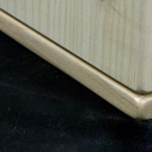 May include: Close-up of the bottom of a wooden table leg with a rounded edge. The leg is made of light wood with a natural finish.