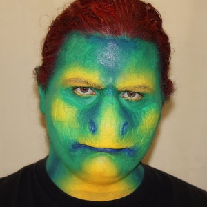 Puede incluir: Una persona con cabello rojo tiene pintura facial verde, azul y amarilla en la cara. La pintura está texturizada para parecer escamas.