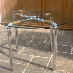May include: A round glass-top table featuring a polished chrome metal frame. The base consists of four tubular legs connected by a central cross-shaped support structure, with tan-coloured accents securing the glass top to the metal frame.