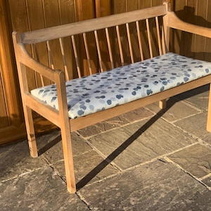 May include: A wooden bench with a light brown finish, featuring a spindle backrest and armrests. The seat is covered in a white cushion patterned with scattered blue and grey circular shapes, providing a flat surface for seating on the furniture.