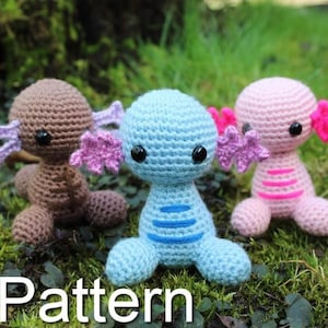 May include: Three crocheted axolotl plushies in different colors: brown, blue, and pink. Each axolotl has pink, crocheted gills and black eyes. The plushies are sitting on a bed of green moss.