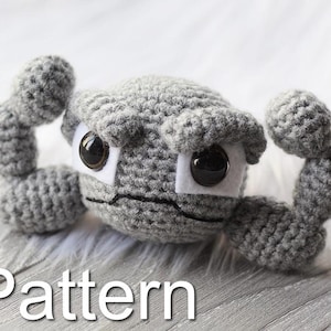 May include: A gray crocheted amigurumi toy with large eyes and a frowning expression. The toy has two large arms raised in a flexing pose. The toy is sitting on a wooden surface with the word "Pattern" written below it.