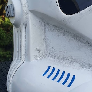 May include: Close-up of a white Stormtrooper helmet with black accents and a dark visor. The helmet features a blue and white striped decal. The helmet has a weathered appearance with gray paint details.