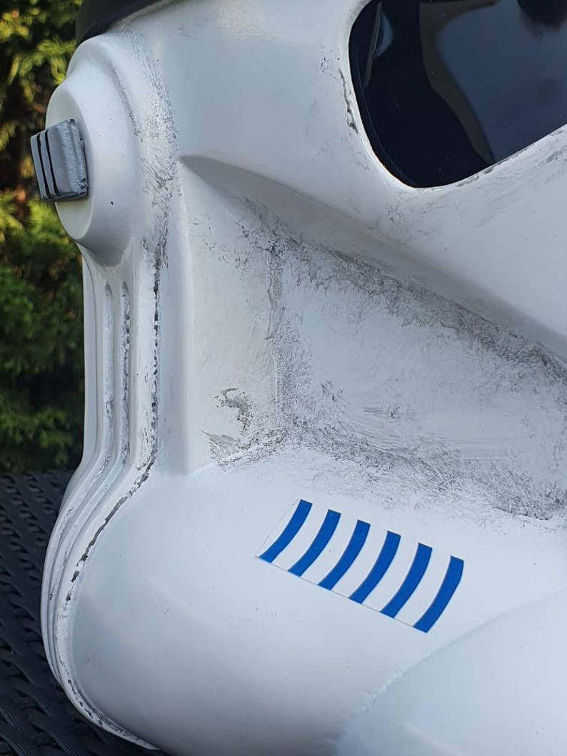 May include: Close-up of a white Stormtrooper helmet with black accents and a dark visor. The helmet features a blue and white striped decal. The helmet has a weathered appearance with gray paint details.