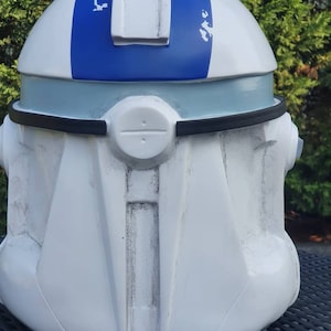 May include: A white and blue helmet with a distinctive design. The helmet features a blue stripe across the top and a light blue band around the middle. The helmet has a black trim and a circular detail on the front. The overall aesthetic suggests a futuristic or sci-fi theme.
