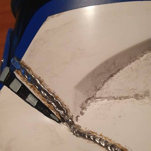May include: Close-up of a white and blue helmet with silver detailing. The helmet has a blue trim and a black section at the top. A silver chain-like design runs along the edge of the helmet.