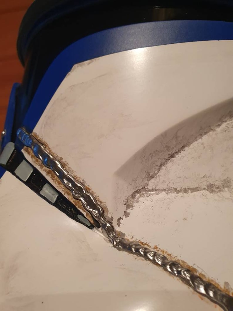 May include: Close-up of a white and blue helmet with silver detailing. The helmet has a blue trim and a black section at the top. A silver chain-like design runs along the edge of the helmet.