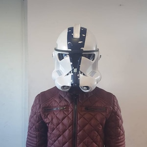 May include: A white and black helmet with a distinctive design, reminiscent of a stormtrooper. The helmet is worn over a burgundy quilted jacket with a zipper closure. The jacket has a diamond pattern.