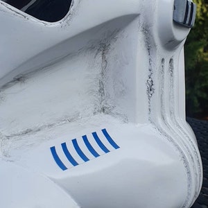 May include: Close-up of a white helmet with black accents, featuring a series of blue stripes on the side. The helmet has a weathered appearance with visible texture and detailing, suggesting a battle-worn aesthetic. The design is reminiscent of a science fiction character.