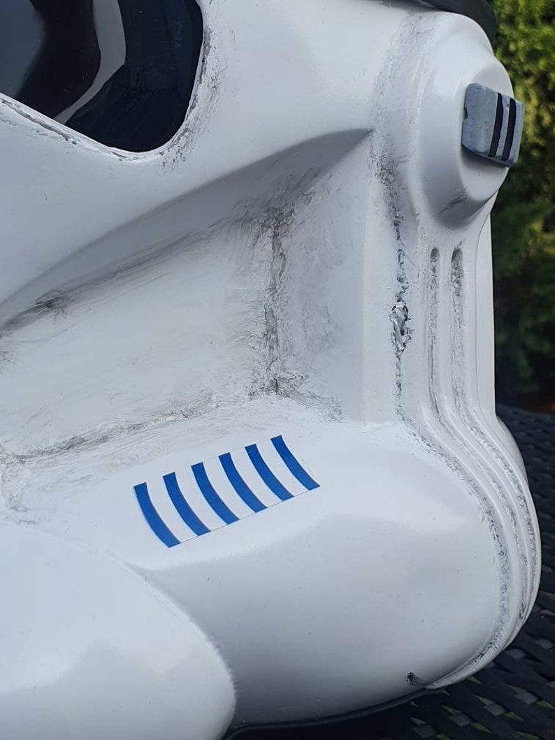 May include: Close-up of a white helmet with black accents, featuring a series of blue stripes on the side. The helmet has a weathered appearance with visible texture and detailing, suggesting a battle-worn aesthetic. The design is reminiscent of a science fiction character.