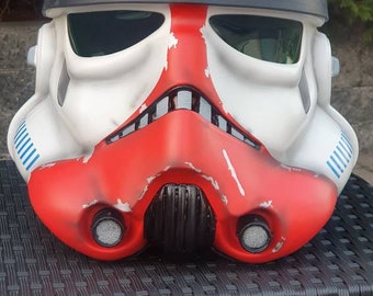 May include: A white and red Stormtrooper helmet with a weathered look. The helmet has a red stripe down the centre and blue stripes on the sides.